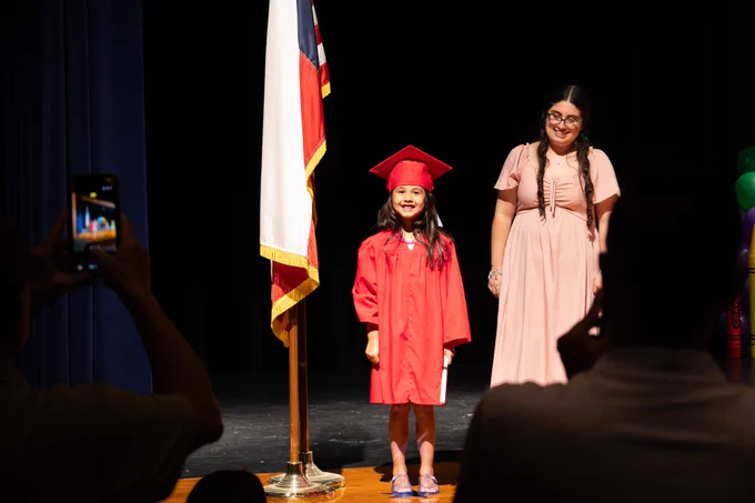 A PreK graduate stands in the spotlight as her photo is being taken by family members cheering her on.