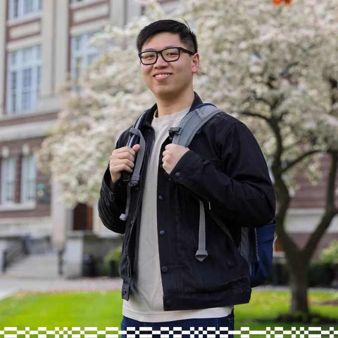 The image shows a person wearing glasses and a backpack, standing in front of a building with flowering trees. The setting appears to be a campus or a place of learning.