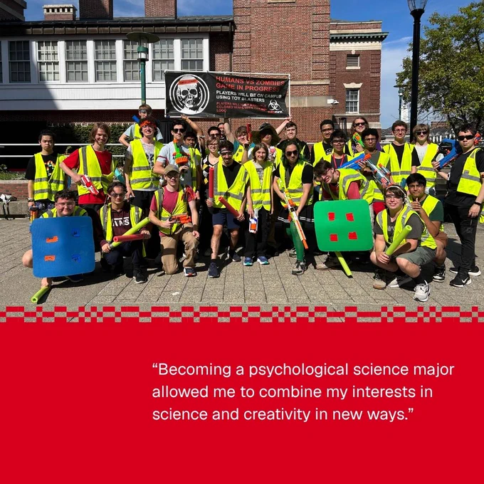 A large group of people wearing bright safety vests and holding colorful shields pose in front of a building with a sign that reads, "Humans vs Zombies Game in Progress." The scene suggests a playful event or game. Below the image, text reads: “Becoming a psychological science major allowed me to combine my interests in science and creativity in new ways.”
