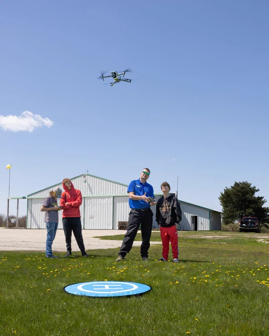 A group of three people is watching a drone flying above a grassy area with a landing pad. A large building and a parked car are visible in the background under a clear blue sky.