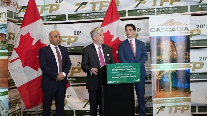 Le ministre Hodgson se tient derrière un pupitre sur lequel figure l'inscription « Building Canada Strong: Strengthening our Forest Sector – Bâtir un Canada fort : Renforcer notre secteur forestier ». Derrière lui se trouvent deux drapeaux canadiens. À sa droite se tient Parm Bains, et à sa gauche, Jake Sawatzky.  