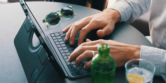 Person typing on a laptop keyboard at a round table with sunglasses and a green bottle nearby.