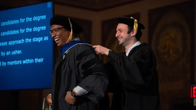A beaming Ph.D. student is hooded by his doctoral advisor.