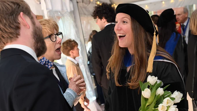 A smiling Ph.D. graduate in regalia holds a bouquet of white tulips and talks with her family.