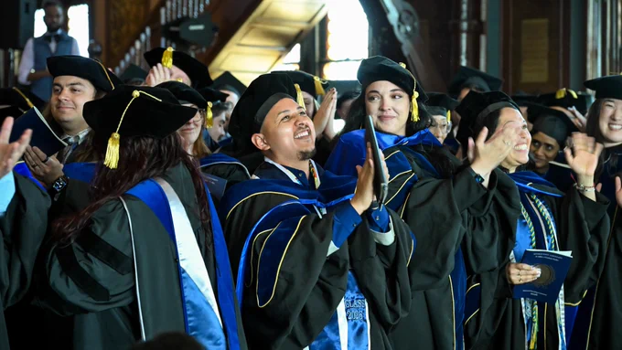 A smiling Ph.D. graduate uplifts his hands in prayer as other graduates appalud around him.