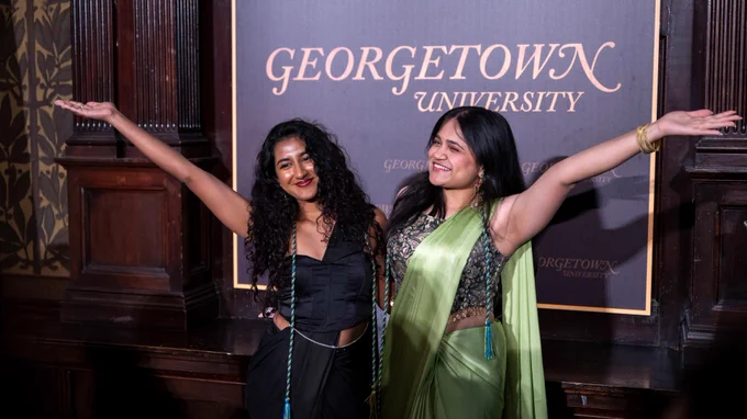 Two female students wearing saris pose together, arms outstretched to frame the Georgetown University logo.