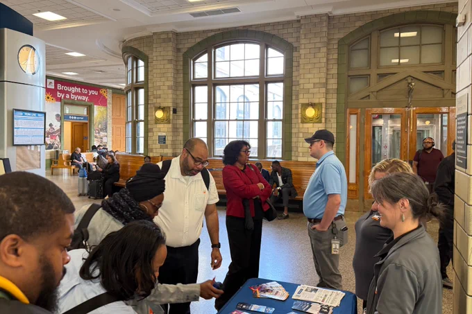 People engaging in conversation and browsing informational materials at a community event inside a historic building with large arched windows.