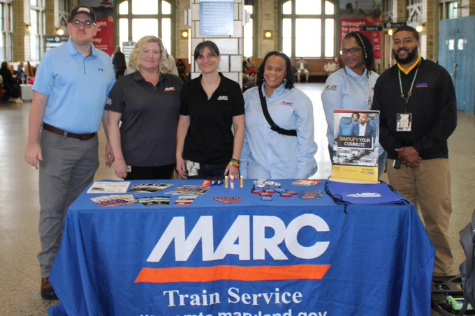 Group of six MARC Train Service representatives standing behind a promotional table with brochures and branded items in a spacious station.