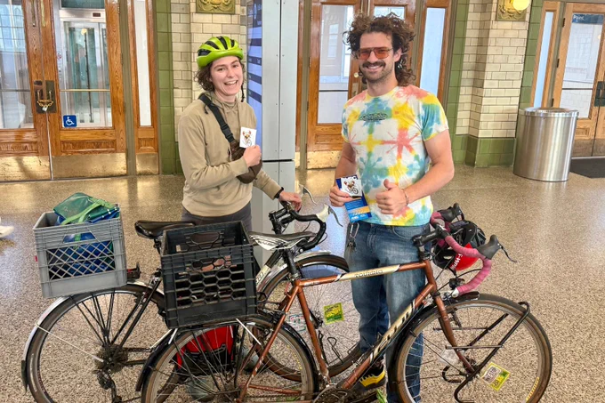 Two cyclists standing indoors with their bikes, one wearing a yellow helmet and beige jacket, the other in a colorful tie-dye shirt giving a thumbs-up.