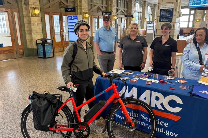Person wearing a helmet stands with a red electric bike inside a train station near a MARC train service information table with staff members behind it.