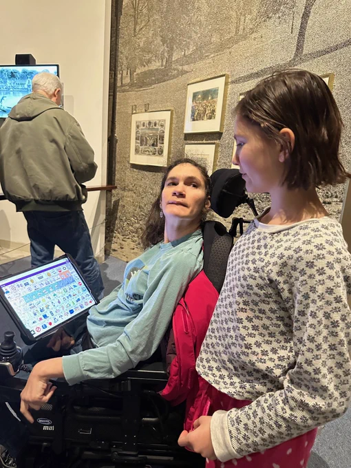 [Image: A person sits in a powerchair with an AAC device in front of them, looking up at a teenager who looks back at them.]