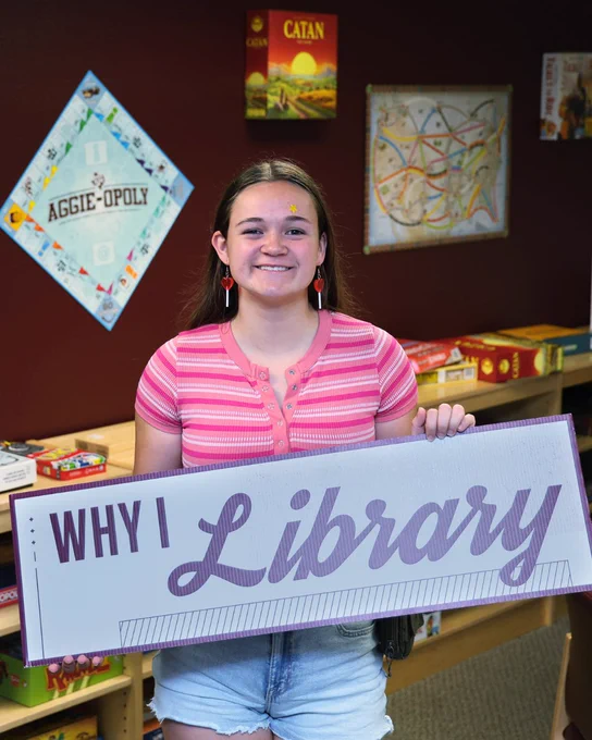 Student holding a "Why I Library" sign. In the background, there are game boards hanging on the wall. 
