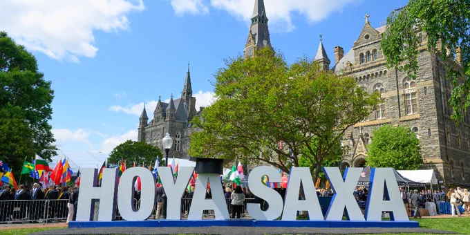 Large white letters spell "HOYA SAXA" in front of Georgetown University’s Healy Hall, surrounded by trees and a clear blue sky. Numerous international flags are visible in the background.