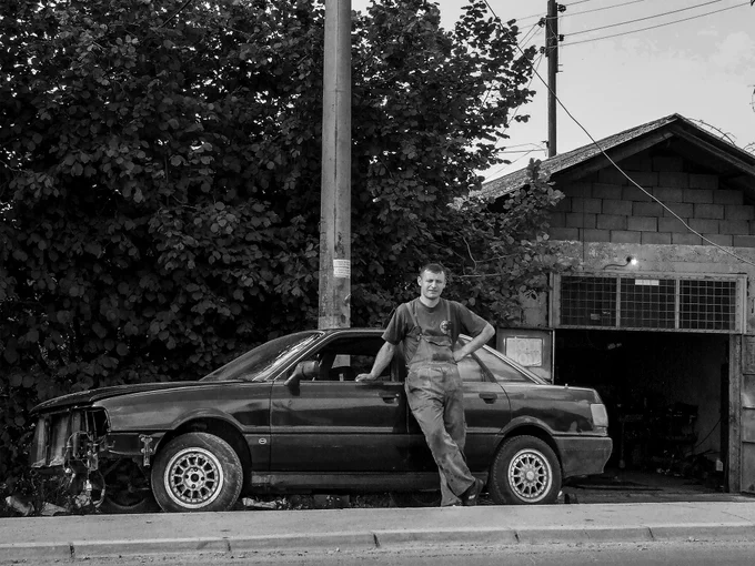 Documentary photograph taken in 2008 in Laplje Selo near Gračanica, Kosovo and Metohija, Serbia. It shows a young auto mechanic standing beside a car outside a garage in a routine working environment. The photograph was made by Serbian photographer Ivan Maksimović.