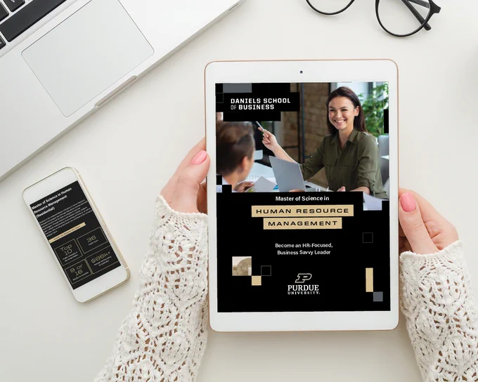 A person is holding a tablet displaying an advertisement for Purdue University’s Master of Science in Human Resource Management program from the Daniels School of Business. The tablet shows an image of two individuals in conversation. Nearby is an open laptop and a smartphone on a desk.