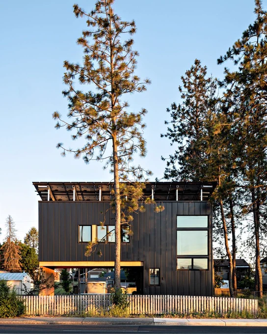 Modern black two-story house with rooftop solar panels, framed by tall pine trees behind a white picket fence.