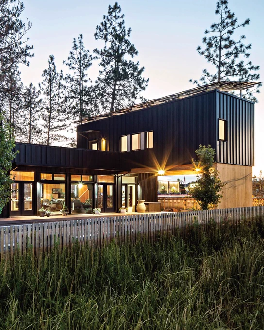 Modern black-clad house with large lit windows and rooftop solar panels, surrounded by pine trees at dusk.