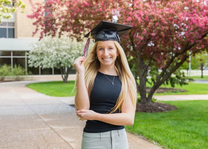 Sienna Rickert wears a black top and gray pants and a graduation cap, standing on a pathway with blossoming trees in the background, smiling and holding a tassel on their cap.