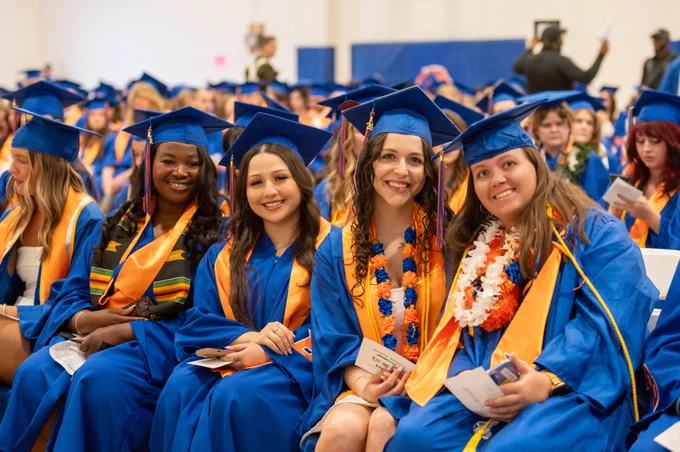 Boise State graduates sitting together and smiling wearing graduation caps and gowns before the Spring 2026 commencement ceremony. 
