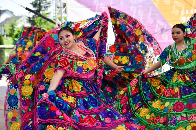 Colorful dancers in vibrant traditional dresses with floral patterns perform energetically at an outdoor event. They gracefully move, holding their wide skirts in flowing arcs. Bright accessories and makeup complete the lively and festive scene.
