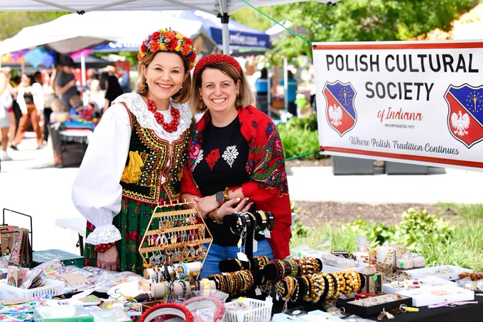 A lively outdoor market booth hosted by the Polish Cultural Society of Indiana. Two individuals in traditional Polish attire stand smiling behind a table filled with various handmade items and jewelry. The booth is decorated with a sign and colorful Polish motifs.