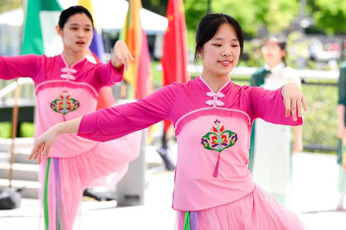 Two performers are dancing, wearing pink traditional outfits with colorful floral embroidery. They are gracefully posed, with flags in the background, set outdoors with greenery visible.