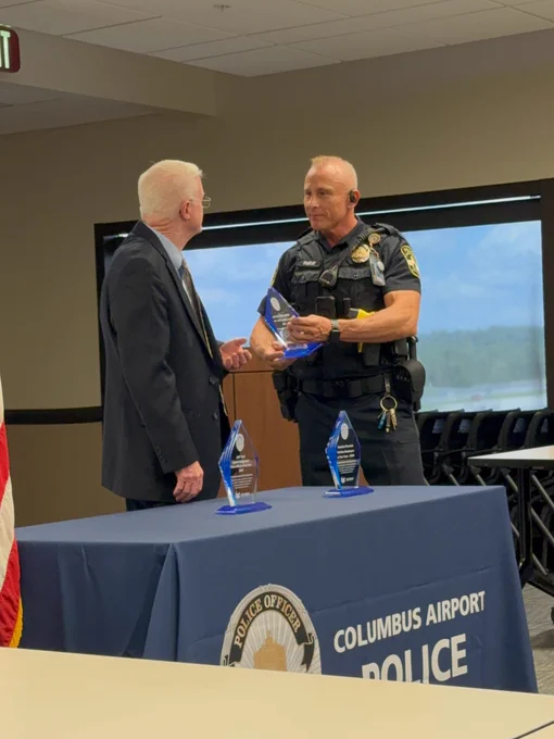 Two individuals are standing at a table with a "Columbus Airport Police" banner. One is in a suit, and the other is in a police uniform. They are holding glass award trophies. An American flag is partially visible on the left.