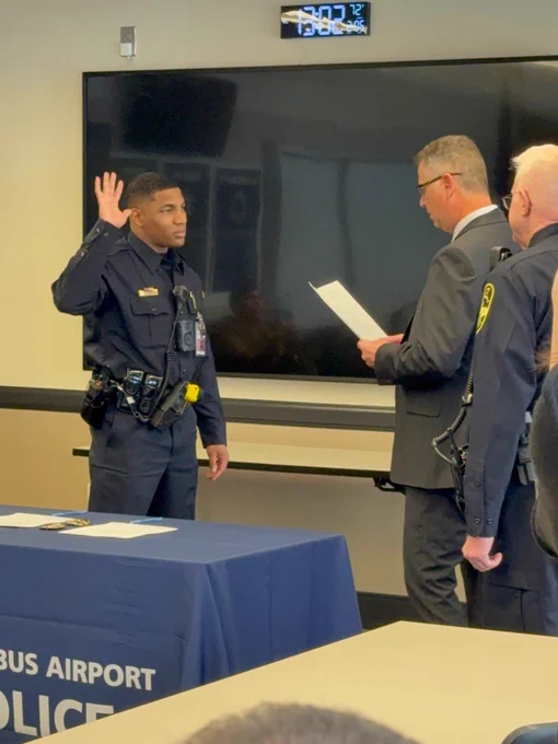 A person in a police uniform is taking an oath with a raised hand, standing in front of a table with a "Columbus Airport Police" banner. Another individual in a suit is reading from a paper. A third person, also in a police uniform, stands nearby. There is a large monitor on the wall behind them.
