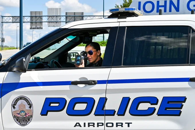A police vehicle labeled "POLICE AIRPORT" is parked with an officer seated inside. The officer is pointing a radar gun outside the window. The background shows part of a sign with the text "JOHN G" visible.