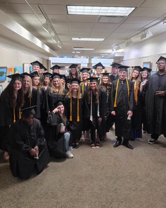 A group of graduates in caps and gowns pose for a photo in a hallway. Some are standing while others are crouching.