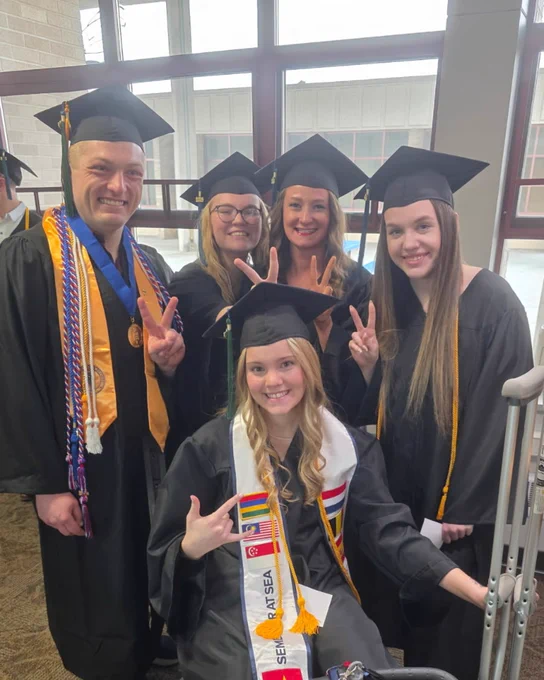 A group of five graduates in caps and gowns are posing indoors. One person is seated on a mobility scooter, wearing a sash with flags. Others are standing behind, smiling, and making peace signs. A window is visible in the background.