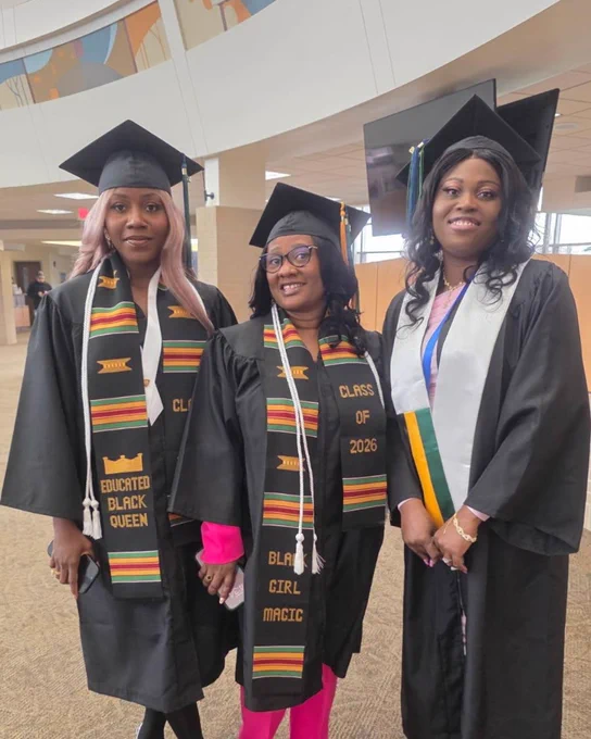 Three individuals in graduation attire stand indoors. They are wearing caps, gowns, and stoles with inscriptions like "Black Girl Magic," "Class of 2023," and "Educated Black Queen." The setting appears to be a hall with large windows and modern decor.
