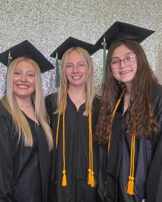 Three people in graduation attire, wearing caps and gowns with gold tassels, stand in front of a sparkly background. They are smiling, celebrating a joyful occasion.