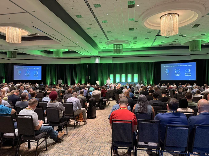 A large conference room filled with attendees sitting in rows, facing a stage with multiple bright screens. The screens display presentation slides with text and graphics. The room is illuminated with green accent lighting.