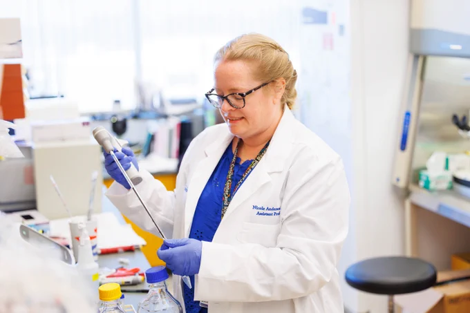 A person wearing a white lab coat and blue gloves is working in a laboratory. They are holding a pipette and standing near a workspace with several laboratory bottles and equipment.