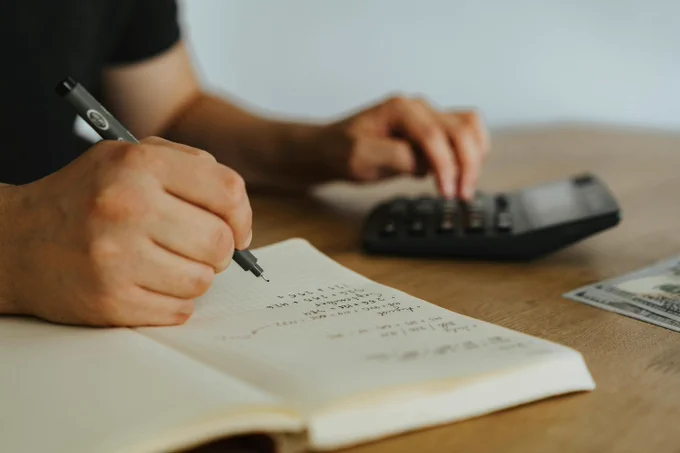A person writes in a notebook while using a calculator, with a U.S. hundred-dollar bill on the wooden table.