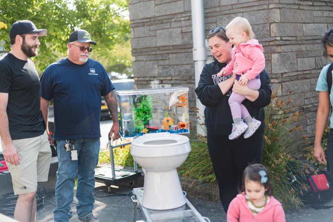 A person holding a child looks on at a toilet with a fish tank for a basin, near City of Bothell employees. 
