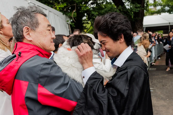 an older man holds up a dog to a younger man in graduation robe