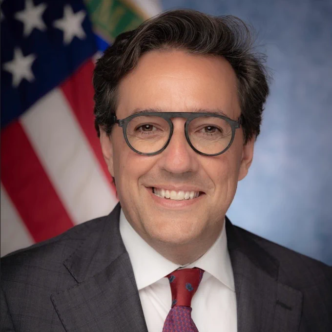 Professional headshot of a man wearing glasses, a dark suit, white shirt, and red patterned tie, smiling in front of a softly blurred background featuring a partially visible American flag and a blue-gray backdrop.