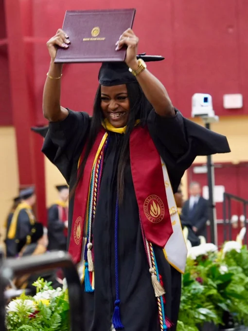 A graduate in cap and gown holds up a diploma with a joyful expression during a ceremony at Rhode Island College, indicated by the logo on the diploma. The background shows other graduates and a decorated stage area.