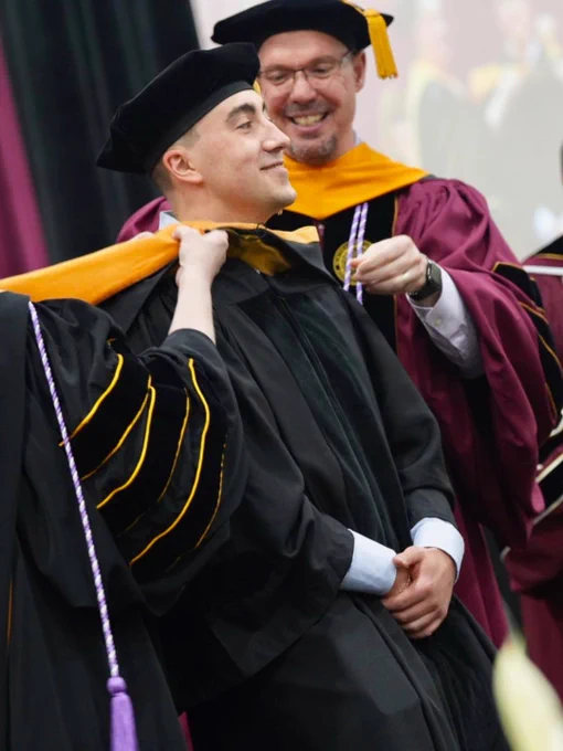 A person is being awarded a doctoral hood during a graduation ceremony. They are wearing a cap and gown, while two individuals in maroon academic regalia perform the hooding. The scene takes place indoors.