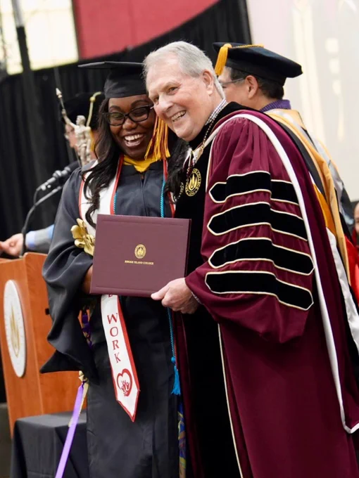 A graduate in a cap and gown stands smiling with a diploma next to a person in academic regalia at a RIC Advanced Degree commencement ceremony.
