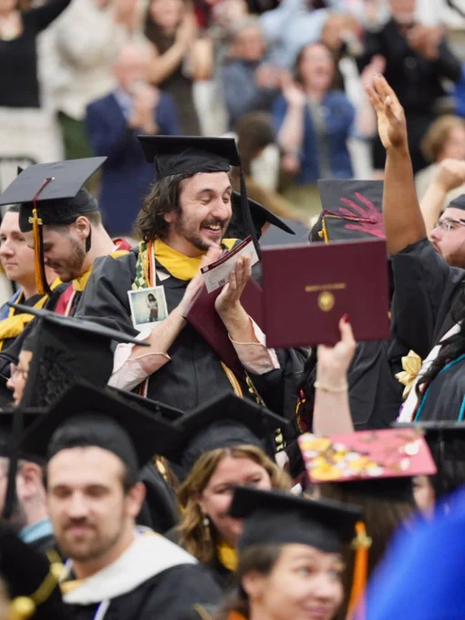 A group of people in graduation attire, featuring black caps and gowns, are gathered together. One person prominently holds a diploma and appears joyful, surrounded by others also in caps and gowns. The background shows an applauding crowd.