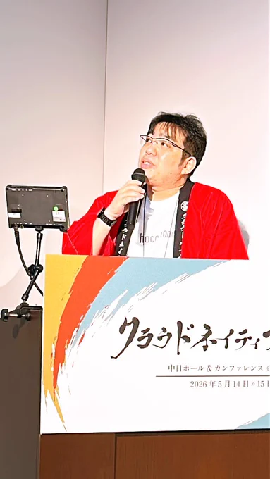 クラウドネイティブ会議と書かれた司会台の奥に赤い法被をきた男性がマイクを片手にお話ししている写真