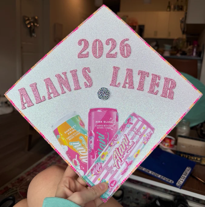 A graduation cap decorated in pink and white, with 3 cans of Alani and the words \"2026 Alanis later\".