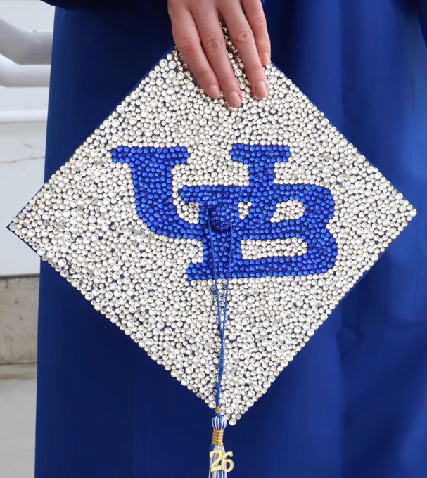 A graduation cap decorated with white rhinestones and blue rhinestones in the shape of the interlocking U B logo.