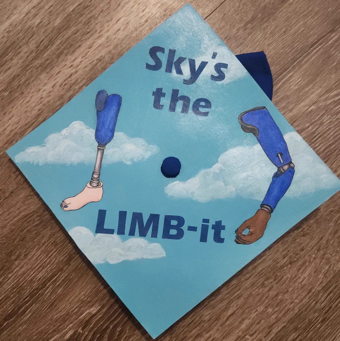 A graduation cap decorated with a cloudy sky, illustrations of a prosthetic arm and leg, and the words \"sky's the limb-it\".