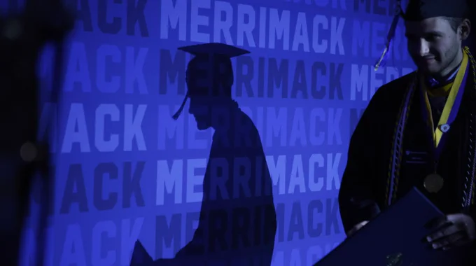 A graduate and their shadow against a Merrimack photo backdrop. 
