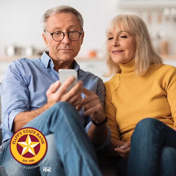 Older couple sitting closely on a couch, looking at a smartphone together in a cozy home setting.