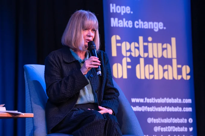 A photo of a woman holding a microphone onstage, speaking to an audience, in front of a big banner with "Festival of Debate" on it